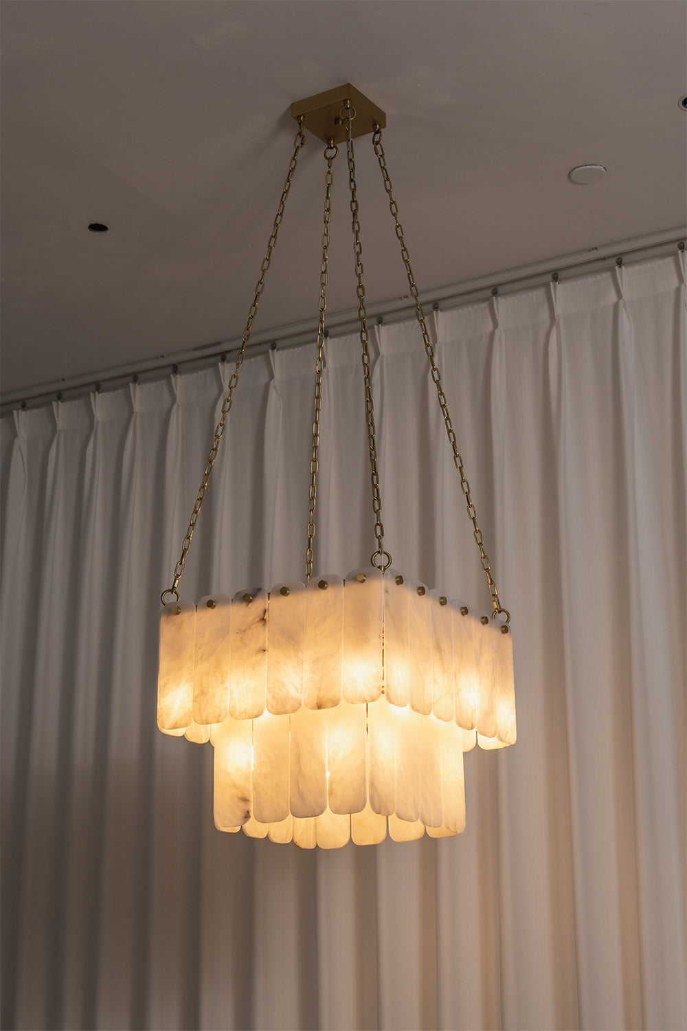 Callista Tiered Alabaster Chandelier | Alabaster, Metal | Studio centered view of the fully lit square tiered alabaster chandelier casting a beautiful warm architectural light against curtains | vexa lamps