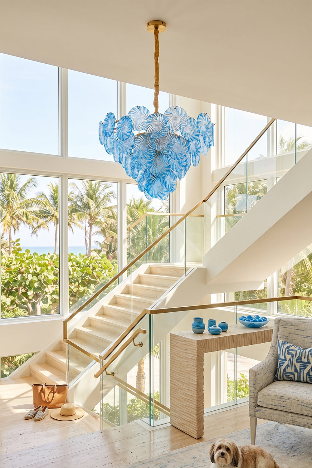 Amara Pink Glass Petal Chandelier | Staircase Foyer Light | Blue glass variant suspended as a grand statement ceiling light near a modern staircase with a scenic beach backdrop | vexa lamps