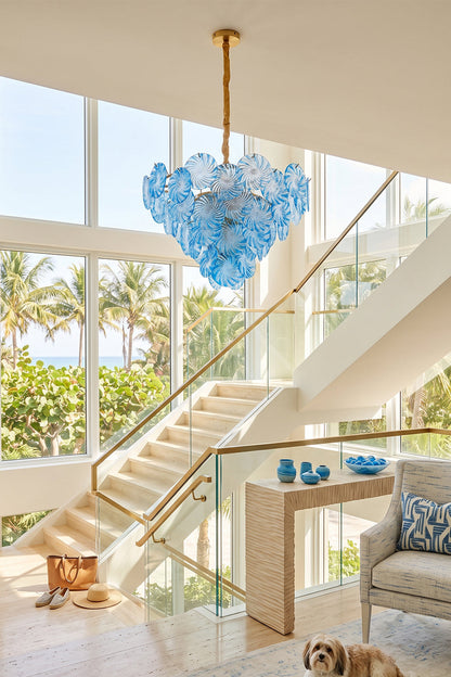 Amara Pink Glass Petal Chandelier | Staircase Foyer Light | Blue glass variant suspended as a grand statement ceiling light near a modern staircase with a scenic beach backdrop | vexa lamps