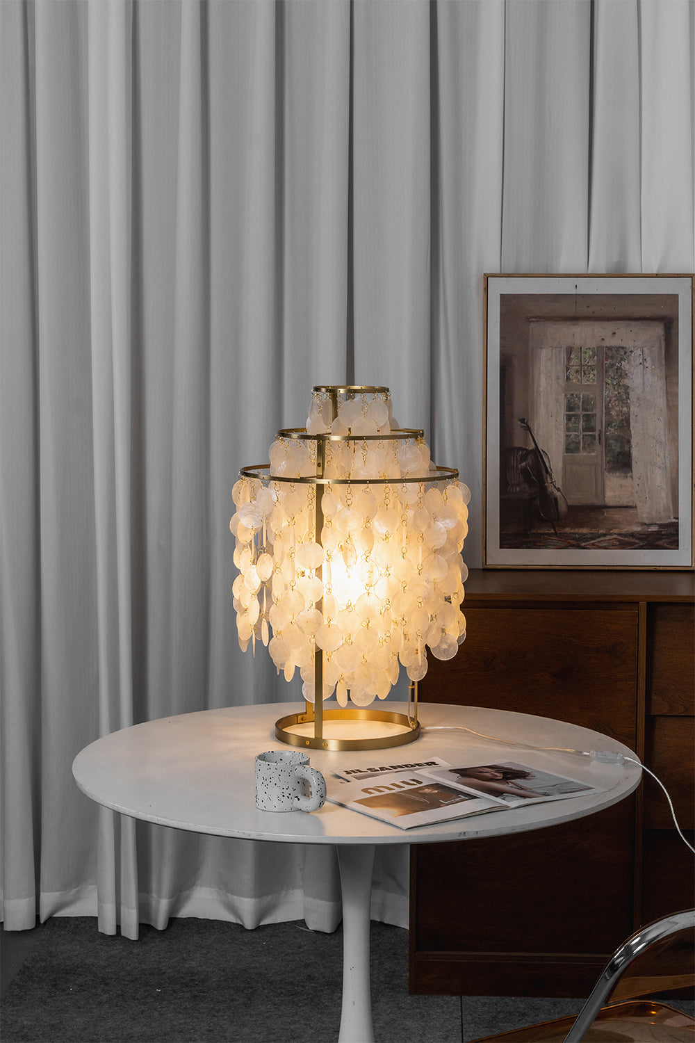 Aurelia Tiered Capiz Table Lamp | Capiz Shell, Metal | Vertical shot of the fully lit cascading capiz shell lamp on a white table against simple white curtains | vexa lamps