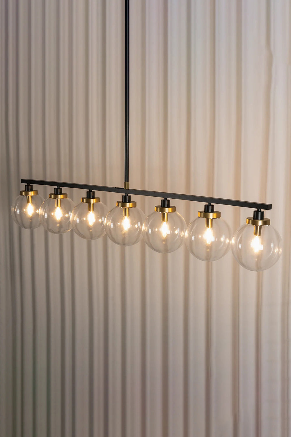 Brooks Linear Glass Globe Chandelier | Glass, Metal | Studio side angle view of the fully lit 7-globe linear suspension bar casting a bright warm light against a pleated white curtain background | vexa lamps