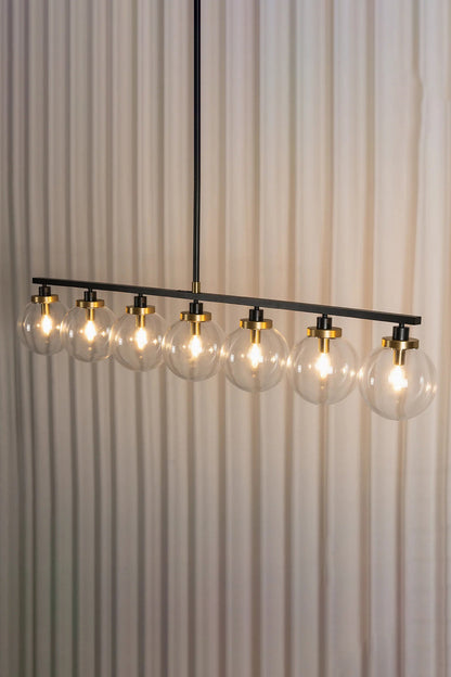 Brooks Linear Glass Globe Chandelier | Glass, Metal | Studio side angle view of the fully lit 7-globe linear suspension bar casting a bright warm light against a pleated white curtain background | vexa lamps