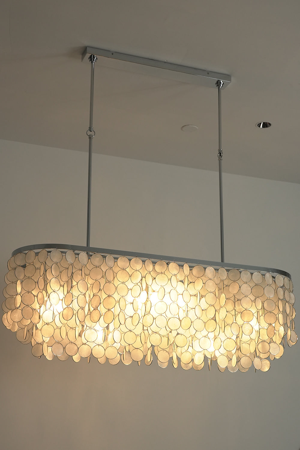 Lit Capiz shell elongated chandelier | Iron | Shell | Low angle view showing warm light filtering through shell discs | vexa lamps