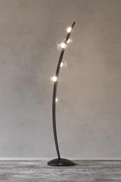 Drosia Dewdrop Floor Lamp | Glass, Metal | Studio view of the fully illuminated minimalist black floor lamp radiating a cozy golden light alignment against a plain grey textured wall | vexa lamps