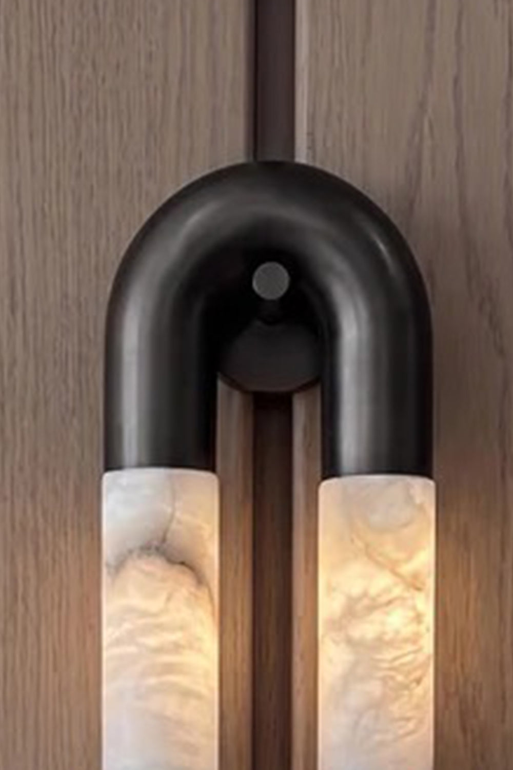 Evadne Alabaster Link Wall Sconce | Alabaster, Metal | Close-up focus on the top curved matte black metallic arch connecting seamlessly to the glowing natural veined alabaster cylinders | Detail shot | vexa lamps