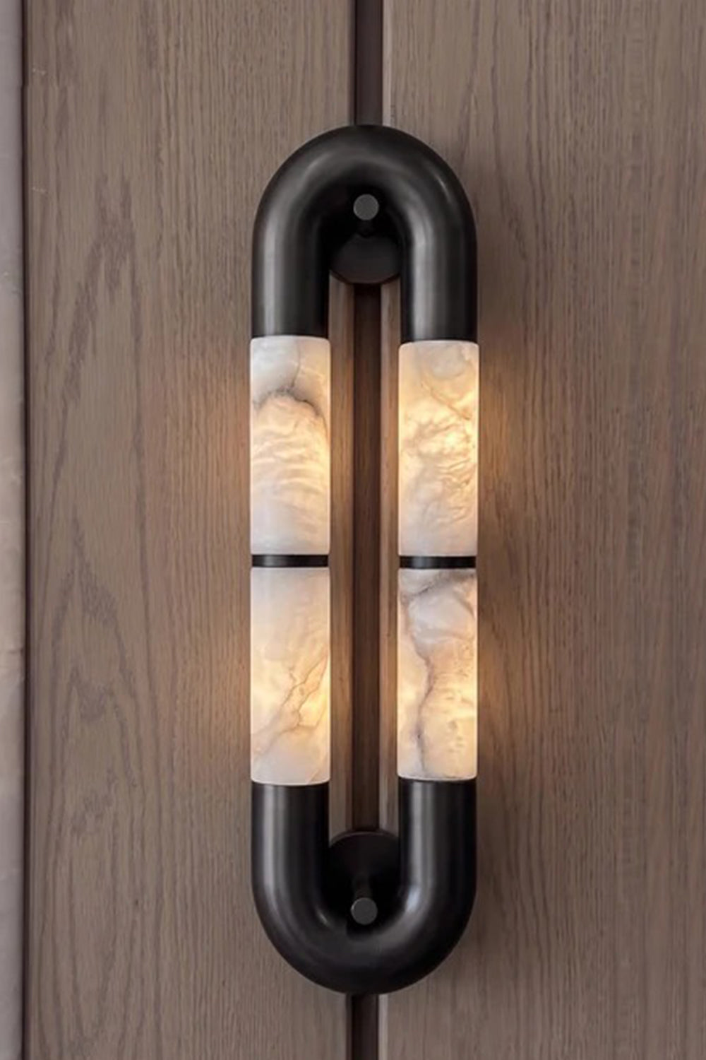 Evadne Alabaster Link Wall Sconce | Alabaster, Metal | Sconce fully lit up on a natural dark wood grain panel background, showcasing the beautiful play of light and shadow | vexa lamps