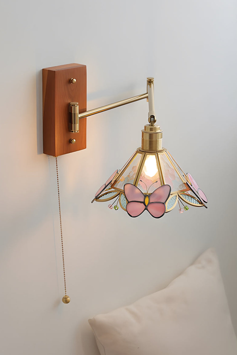 Iris Butterfly Stained Glass Wall Lamp | Stained Glass, Metal, Wood | Mounted on a plain white wall above a soft cream pillow, showcasing the full extension of the gold brass swing arm and the hanging pull chain | vexa lamps