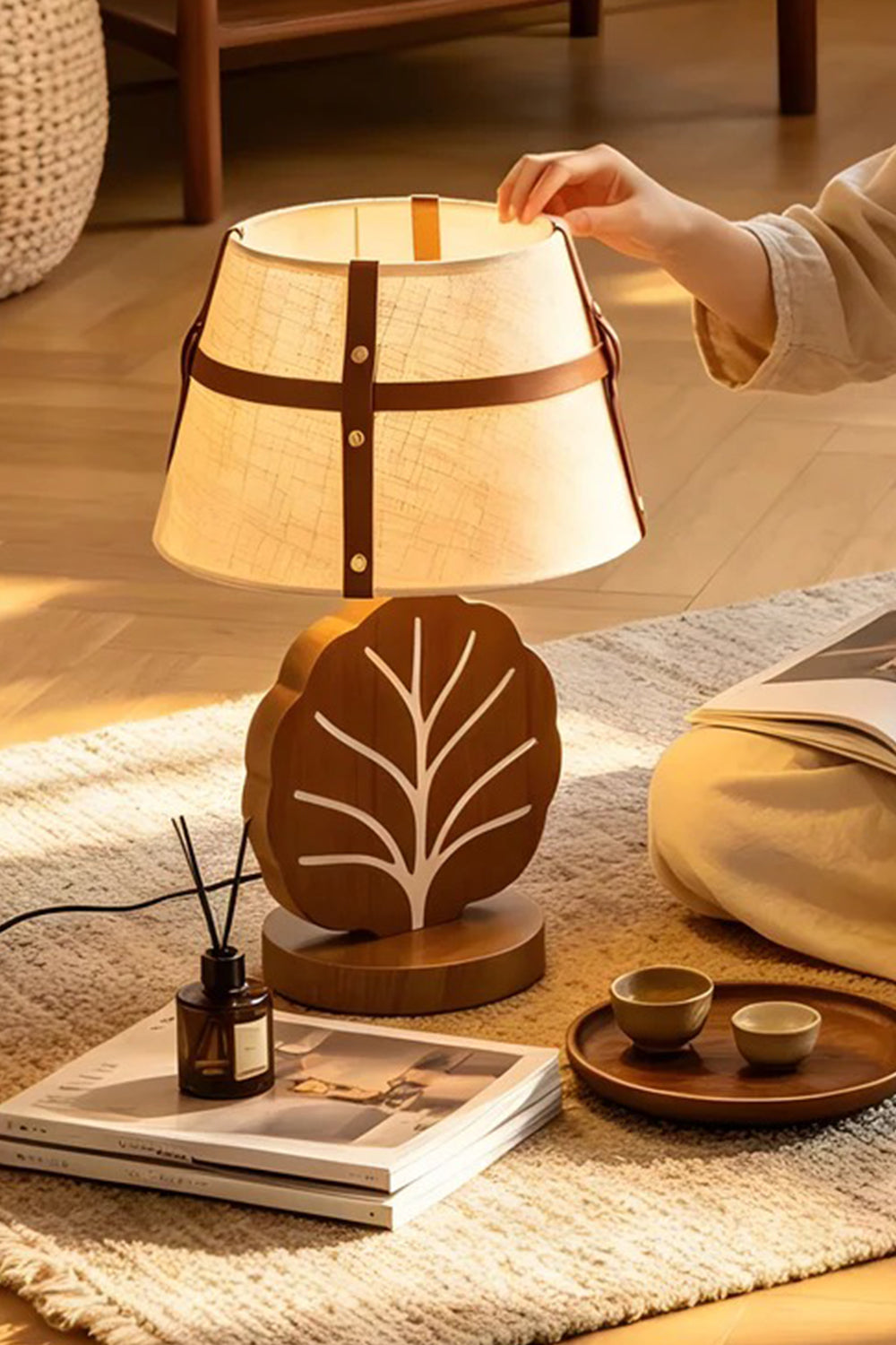 Leafora Botanical Leaf Table Light | Wood, Fabric, Leather | Cozy indoor scene on a textured floor rug next to books, showing a hand interacting with the fully illuminated leaf table lamp | vexa lamps