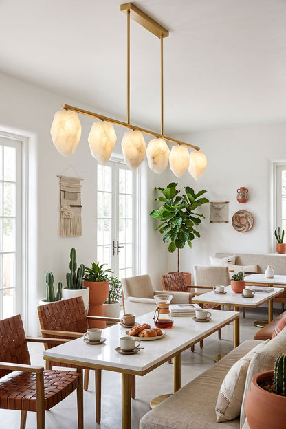 Manon Alabaster Drop Linear Chandelier | Natural Alabaster and Brass | Hanging above a wooden dining table in a bright room with potted plants | vexa lamps