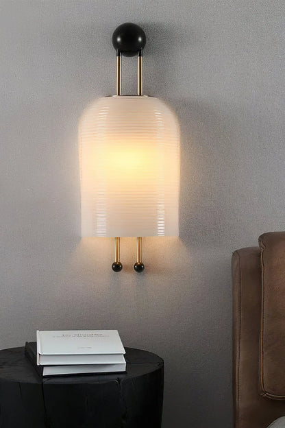 Marlow Linear Ribbed Glass Wall Lamp | Glass, Metal | Close-up shot of the lit ribbed sconce on a grey textured wall casting a warm glow over a black nightstand with a book | vexa lamps
