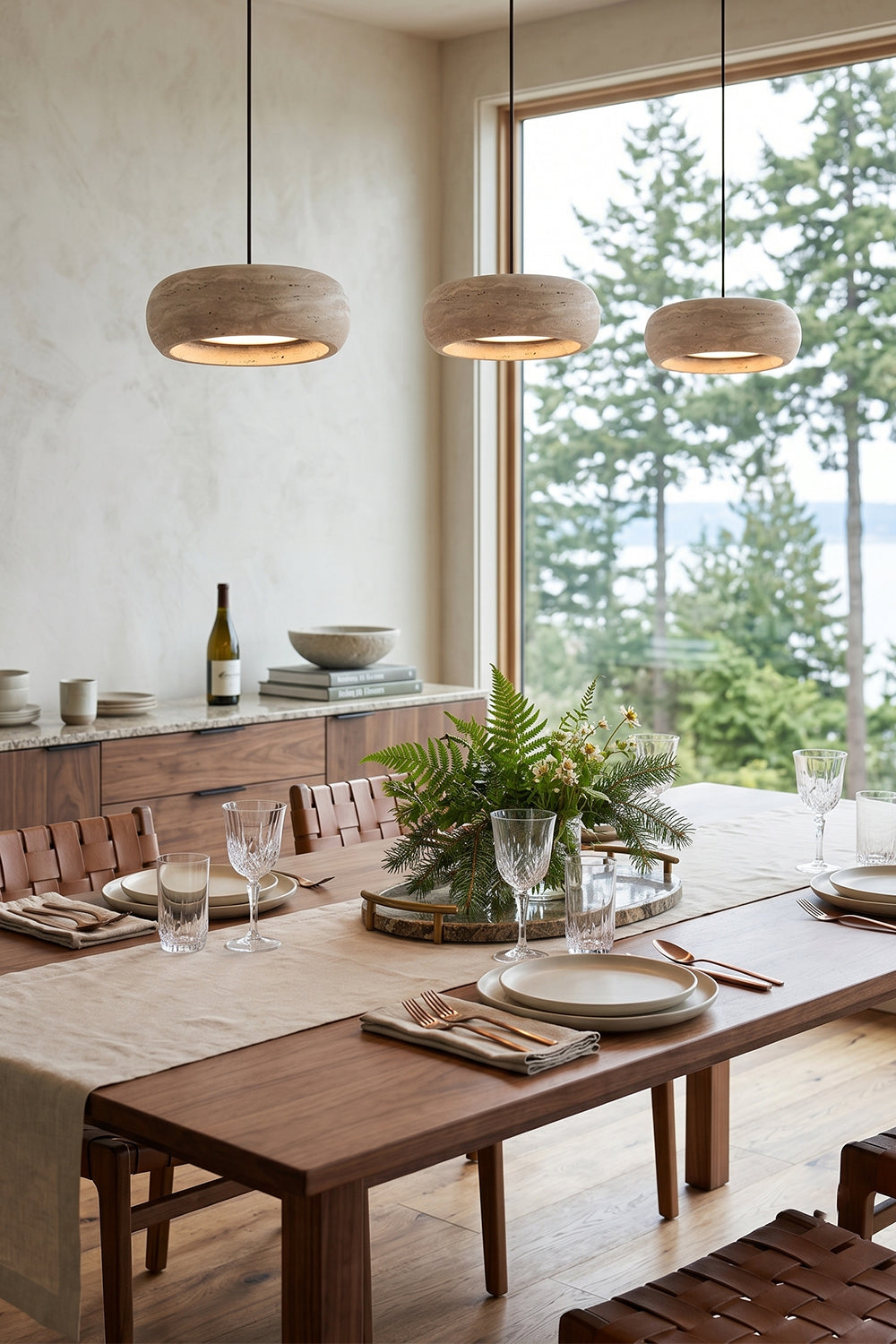 Maro Travertine Donut Pendant Light | Travertine, Metal | Three linear stone pendant fixtures hung above a contemporary wood dining table in a sunlit open-concept home interior | vexa lamps