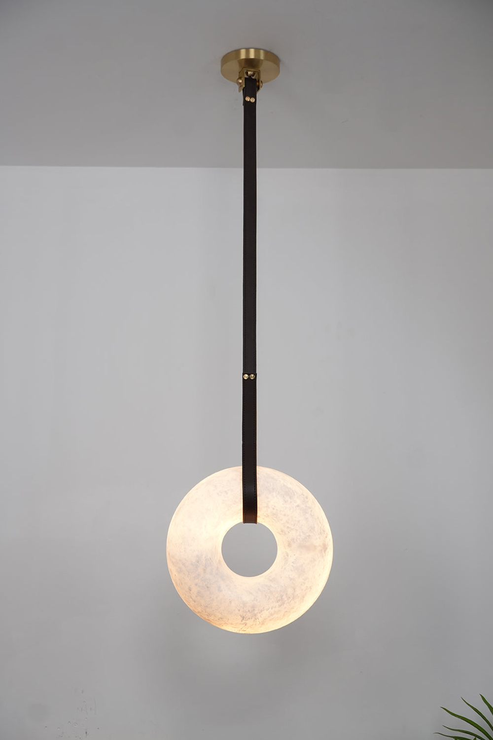 Nerissa Alabaster Ring Leather Pendant Light | Natural Alabaster, Leather and Brass | Single-strap disc variant illuminated softly on a grey wall | vexa lamps