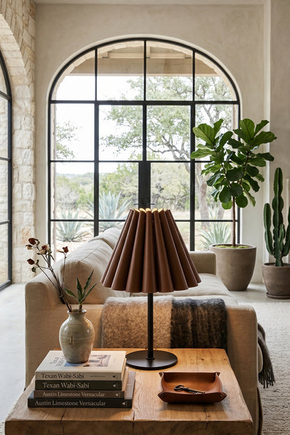 Rowan Leather Pleated Table Lamp | Leather, Metal | Positioned on a wooden coffee table in a modern lounge space featuring a large black framed arched window | vexa lamps