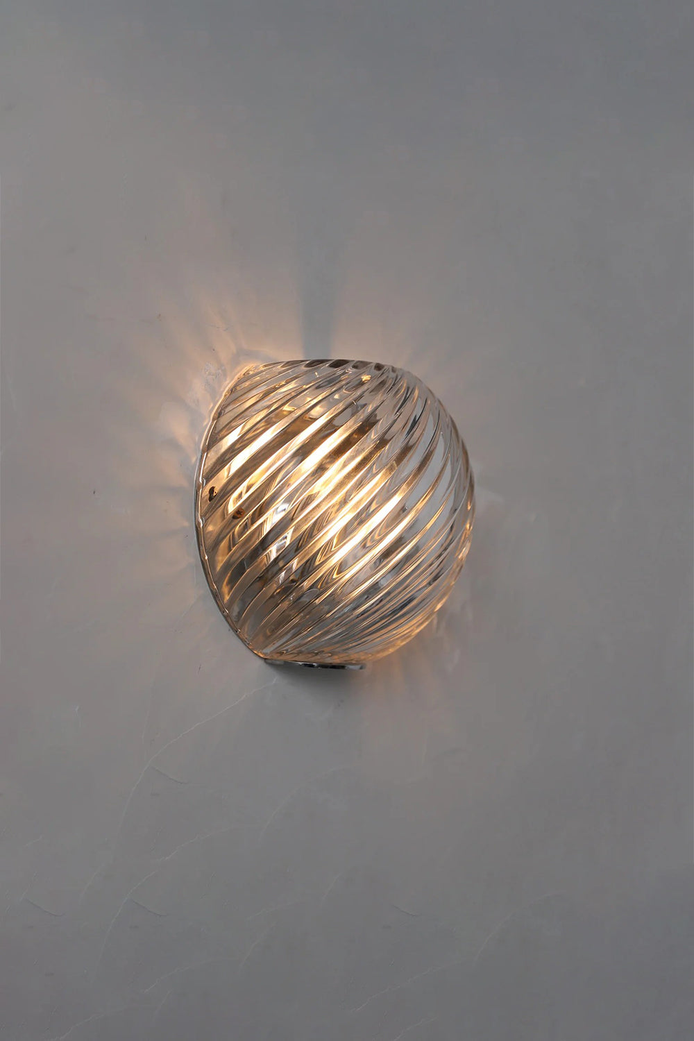 Silas Ribbed Glass Wall Sconce | Ribbed Glass, Metal | Staged lit studio view of a clear glass variant casting beautiful swirled ribbed light patterns against a grey wall | vexa lamps