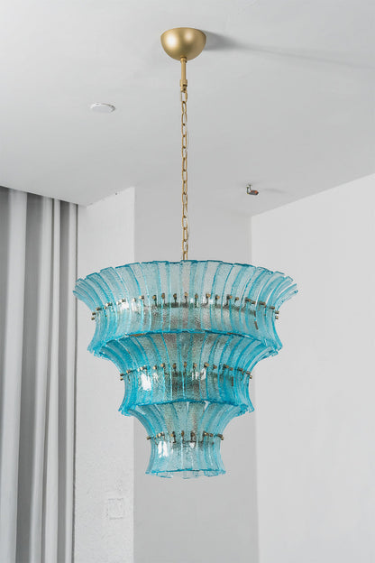 Thalassa Aquamarine Textured Glass Chandelier | Glass, Metal | Studio staged view of the tall multi-tiered lit aquamarine glass chandelier casting a rich sea-colored glow near grey window curtains | vexa lamps