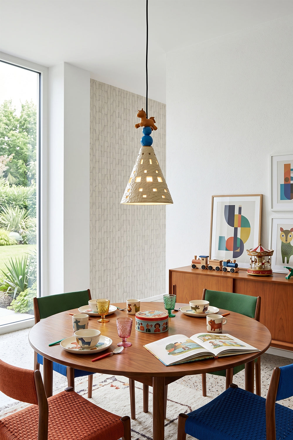 Whimsy Ceramic & Resin Pendant Light hanging above a stylish dining table with colorful chairs, books, and tableware.